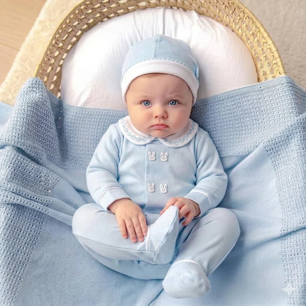 Bebê deitado em berço, vestido com macacão azul claro e touca, coberto por manta azul, roupa delicada para recém-nascido.