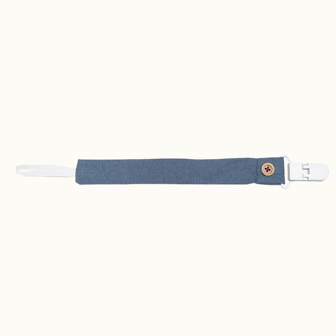Suporte de chupeta em ganga azul claro com clip branco e botão decorativo, fundo neutro.