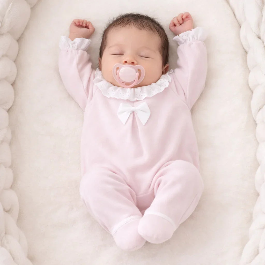 Bebê dormindo em ninho, vestindo babygrow rosa com laço e gola de babados delicada.