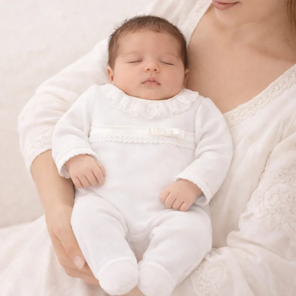 Bebê dormindo no colo, vestindo babygrow branco com detalhes delicados em renda