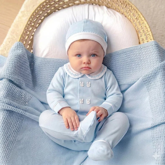 Bebê deitado em berço, vestido com macacão azul claro e touca, coberto por manta azul, roupa delicada para recém-nascido.