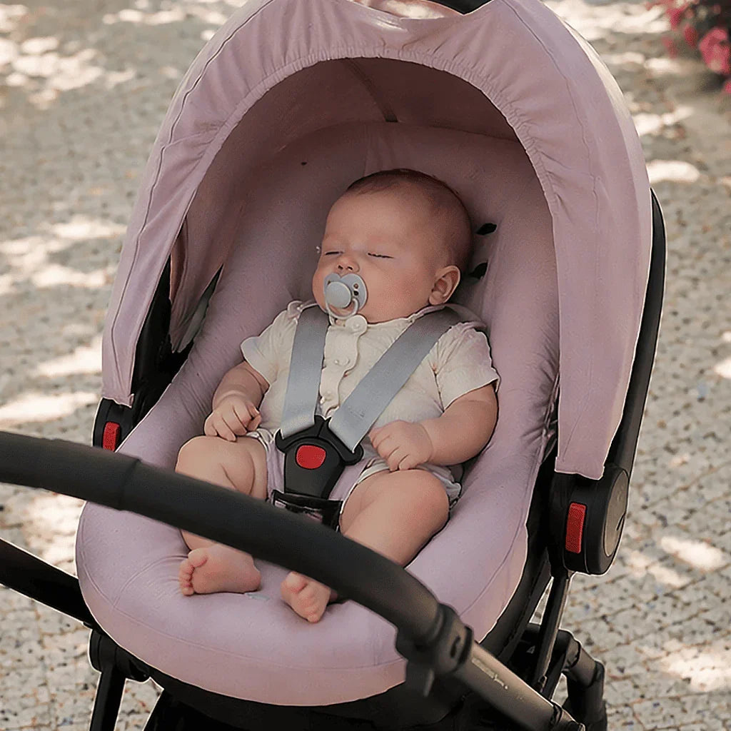 Bebê dormindo em cadeirinha rosa no carrinho, usando roupa confortável ao ar livre