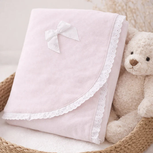 Manta de veludo rosa para bebé com laço branco e bordado, em cesto ao lado de ursinho de peluche.