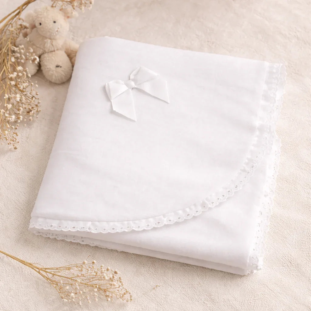 Manta de veludo branca para bebê, com laço decorativo e acabamento rendado, sobre superfície bege.