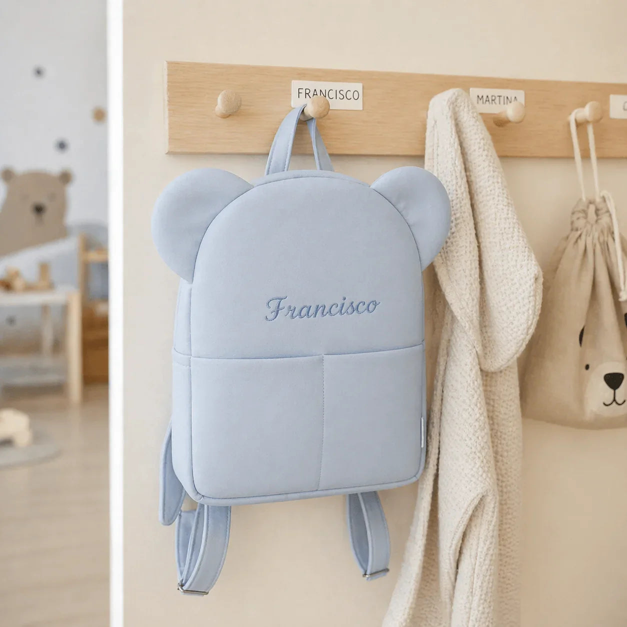 Mochila infantil azul com orelhas, pendurada em cabideiro de creche, ambiente decorado para crianças.
