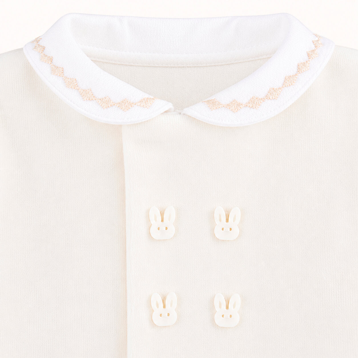 Detalhe de babygrow bege para bebê com gola bordada e botões decorativos de coelhinho.