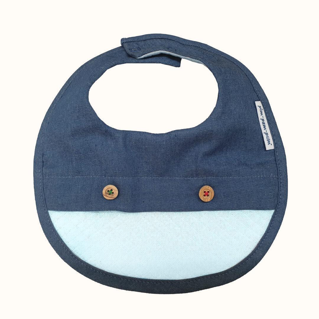 Babete impermeável azul denim com botões decorativos para bebé, fundo claro.