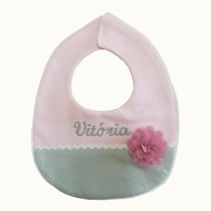Babete de malha rosa e verde para bebé, com flor decorativa e nome 'Vitória' bordado.