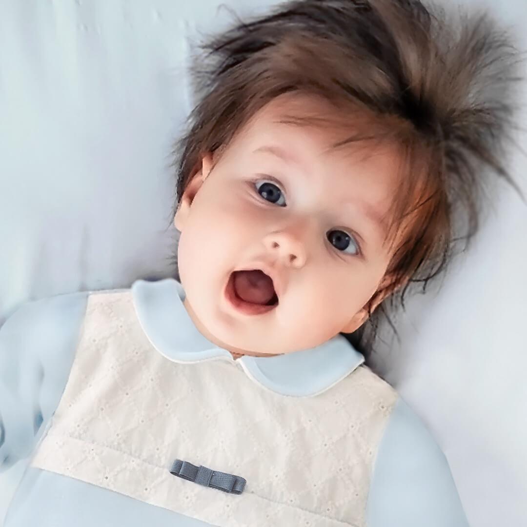 bebê deitado usando babygrow azul com detalhe em renda branca e laço, roupa de bebê confortável