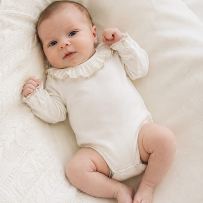 Bebé vestido com um body com gola folho e renda bege em malha algodão bege, sem etiquetas irritantes no decote e abrir de cima a baixo para facilitar a muda da roupa