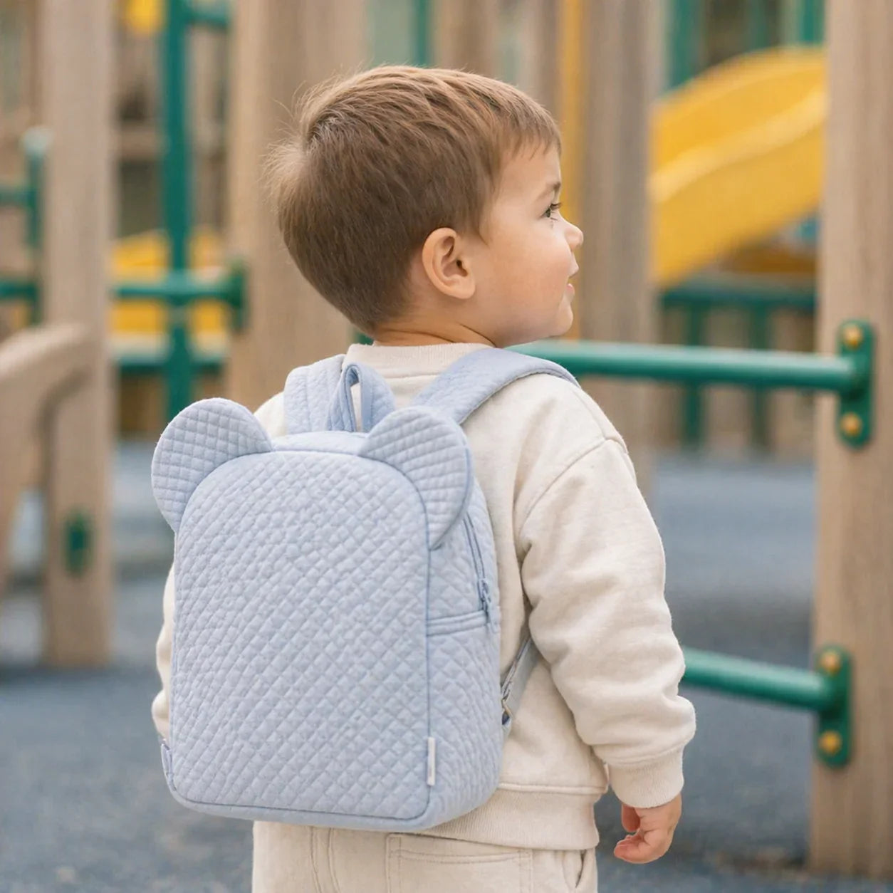 bebé com mochila azul acolchoada com orelhas em parque infantil, roupa confortável de bebé.