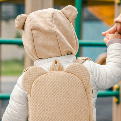 Bebé de casaco branco com mochila e touca bege de orelhinhas em parque ao ar livre.