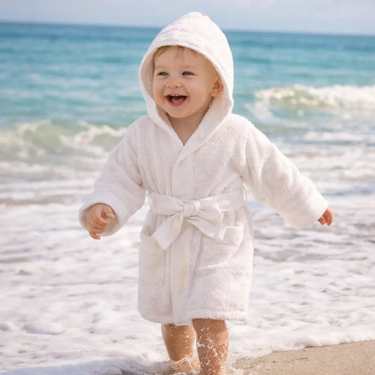 bebê sorrindo de roupão de algodão branco na praia, roupa confortável para bebê