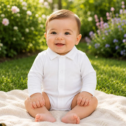 Bebé sorridente vestido com body tipo camisa branco algodão confortável, sentado na grama com flores ao fundo