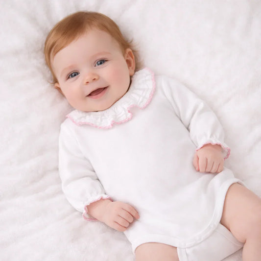 Bebé sorrindo usando body branco com gola franzida e detalhes rosa, deitado em manta macia.