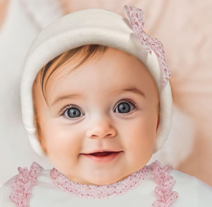 Bebê sorrindo com gorro branco e laço rosa, roupa delicada para recém-nascido.