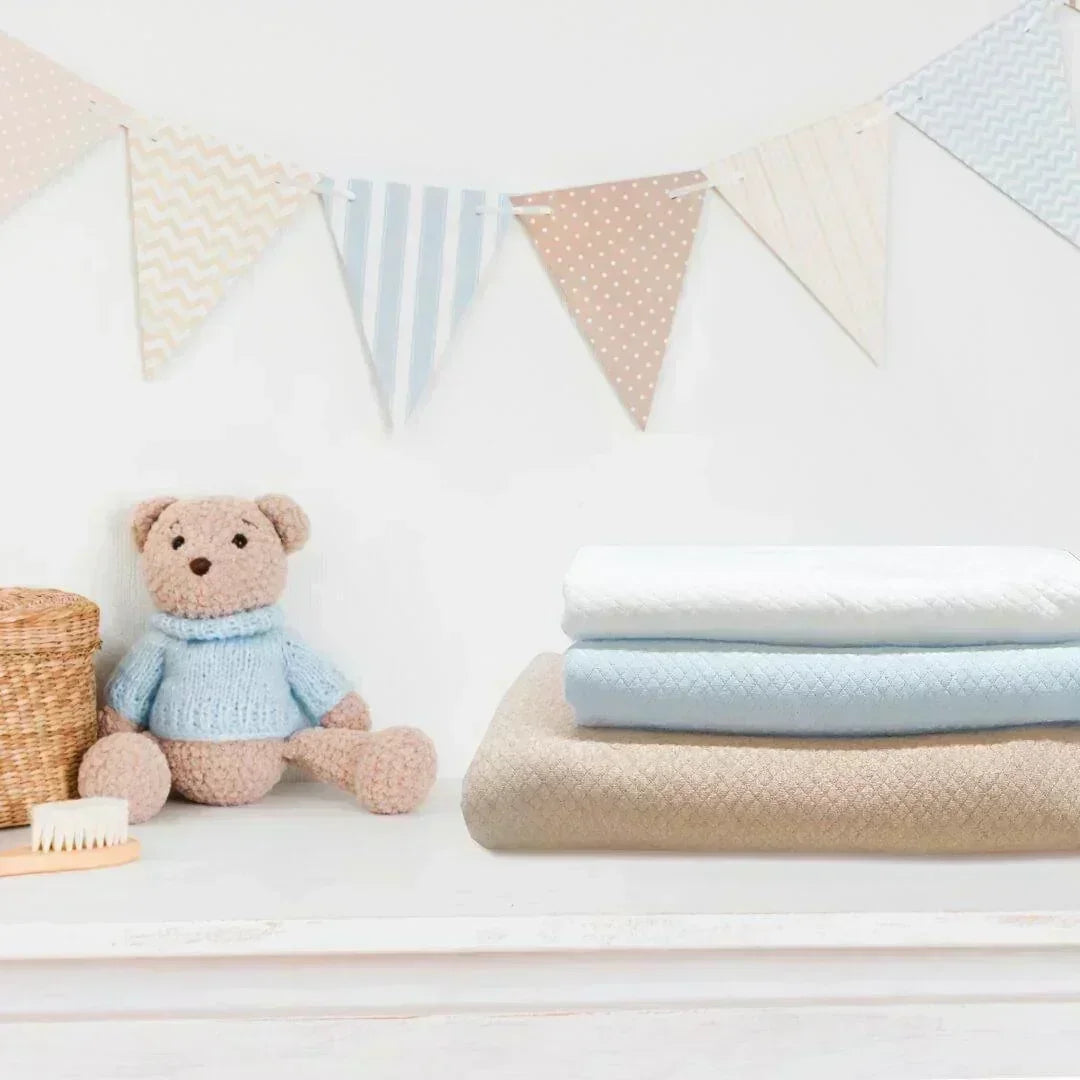 Trio de mantas para bebê em tons camel, azul e branco, ao lado de ursinho de pelúcia.