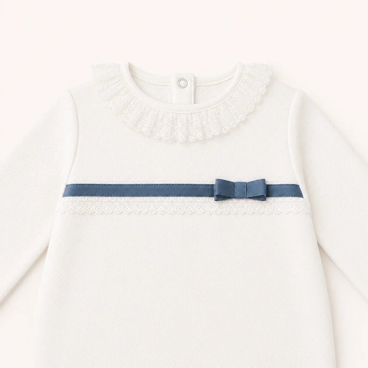 Pormenor de babygrow bege com renda e laço azul, ideal para enxoval confortável e delicado.