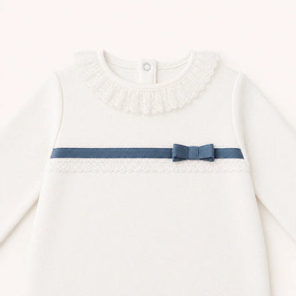 Pormenor de babygrow bege com renda e laço azul, ideal para enxoval confortável e delicado.
