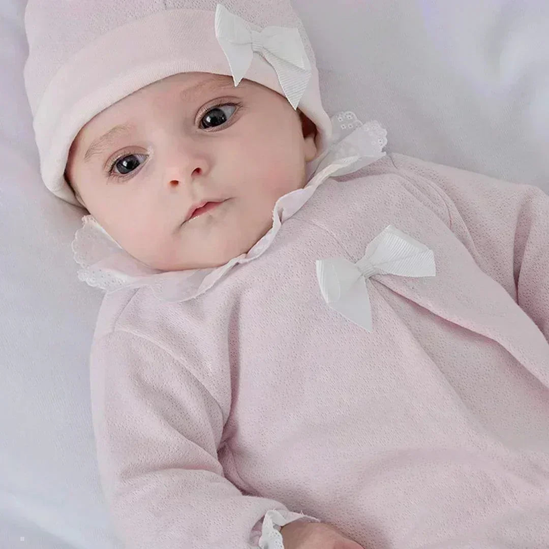 Bebê usando macacão rosa claro de algodão com laço branco e touca combinando, deitado em fundo branco.